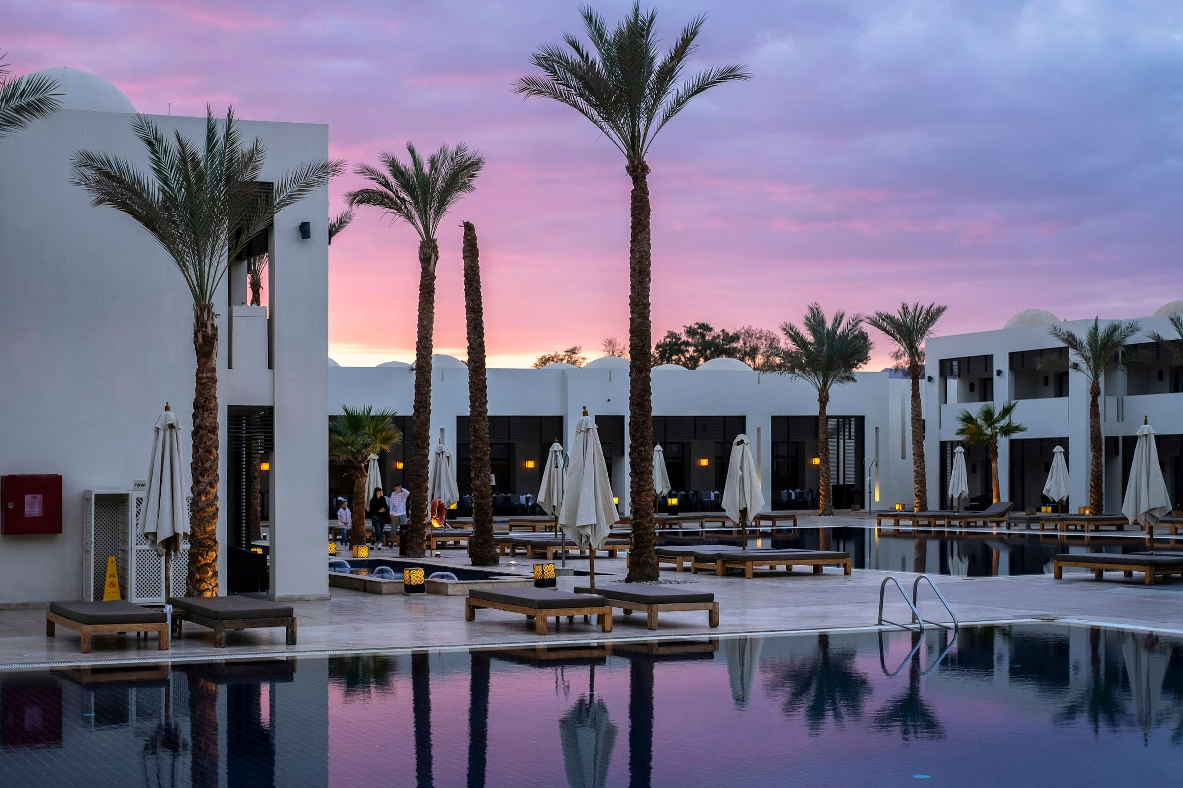 A white resort with tall palms reflected in a pool at dusk